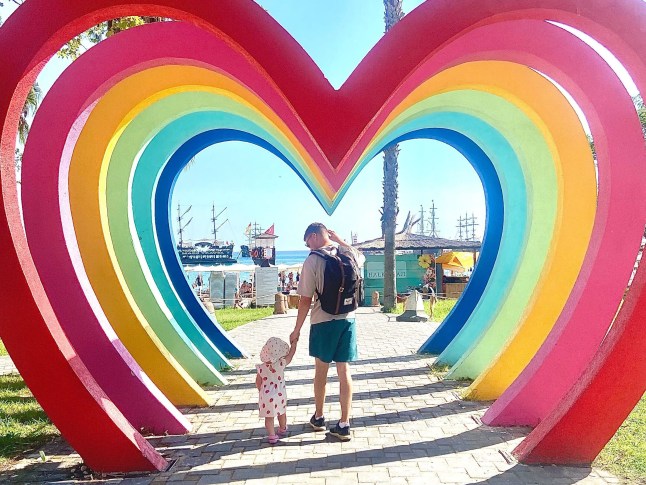 A man walks hand in hand with his small daughter under a rainbow heart structure