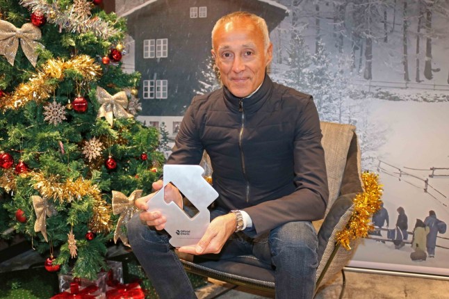 Andrew Ridgeley sat next to a Christmas tree holding his Christmas number one single award. He is wearing blue denim jeans and a grey jumper